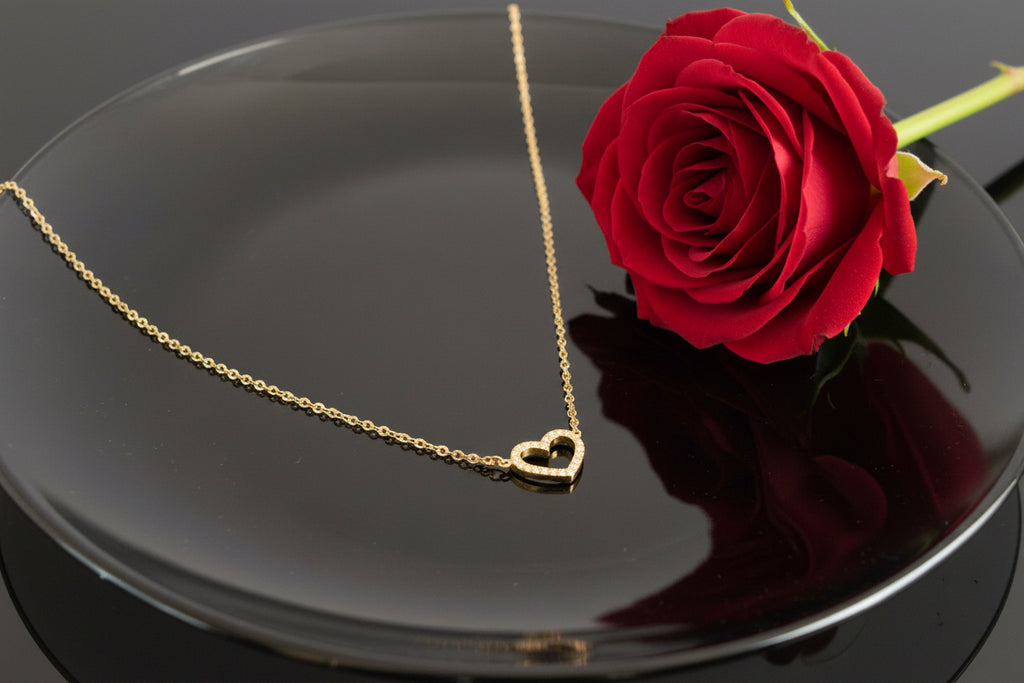 Eine vergoldete kette mit herzanhänger liegt auf einem dunklen Teller daneben liegt eine Rose

